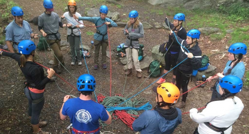 A group of people wearing safety gear stand in a circle and are connected by ropes, likely in an exercise. 